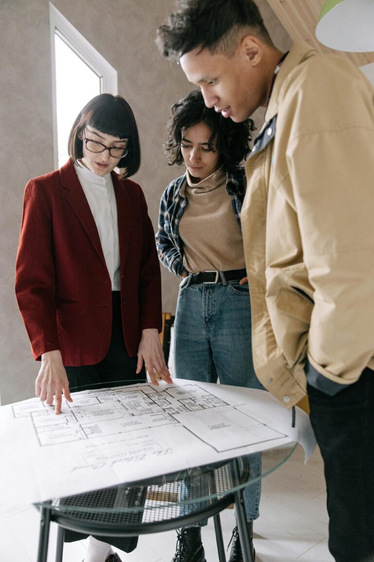 A real estate agent discussing blueprints with a young couple at a glass table indoors.